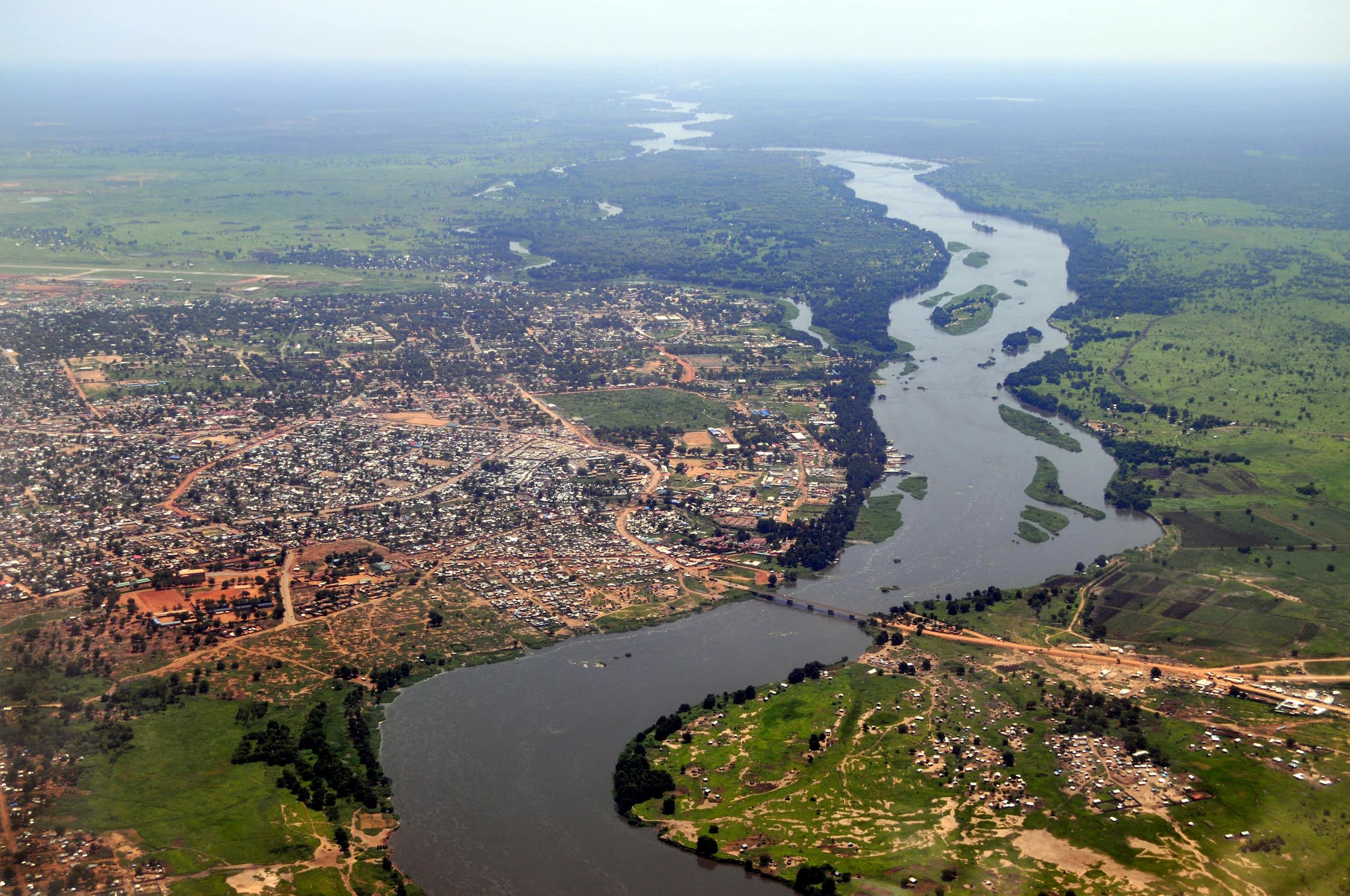 An aerial photograph of a river wending its way through green valleys, flanked on the left of the image by a sprawling city