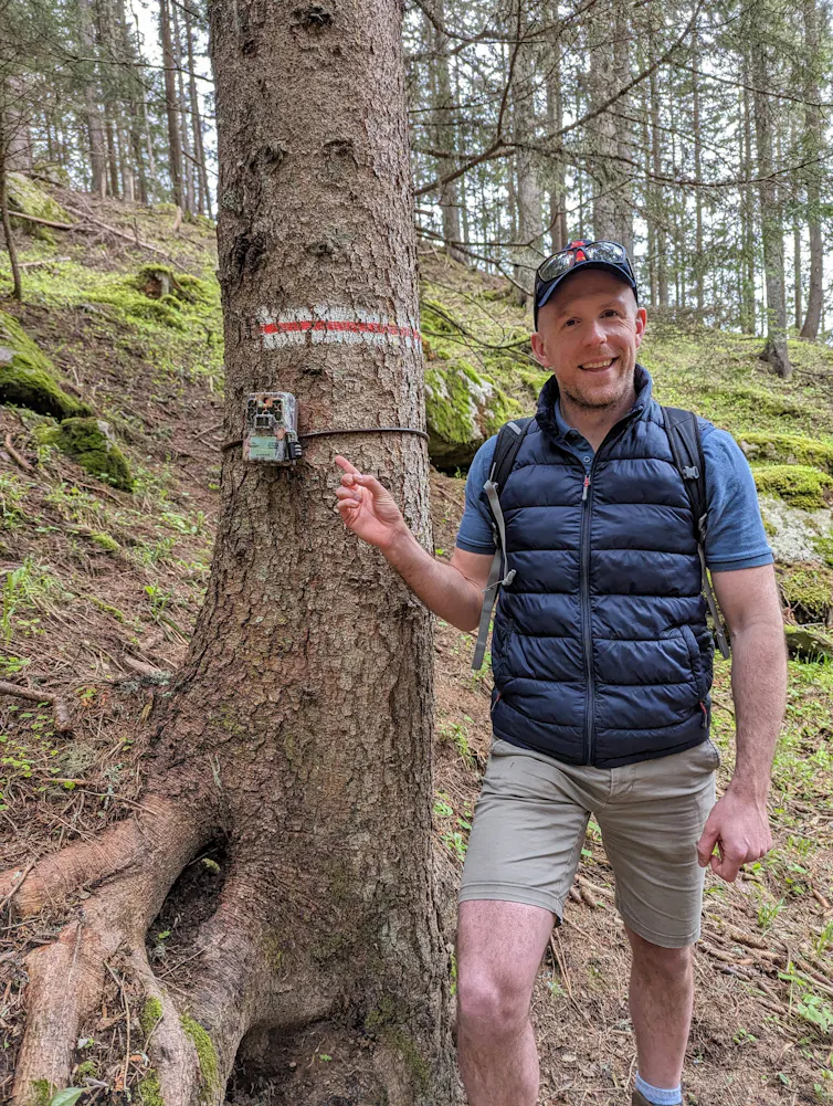 Man in navy coat and grey shorts standing by tree with camera strapped around trunk, green forest in background