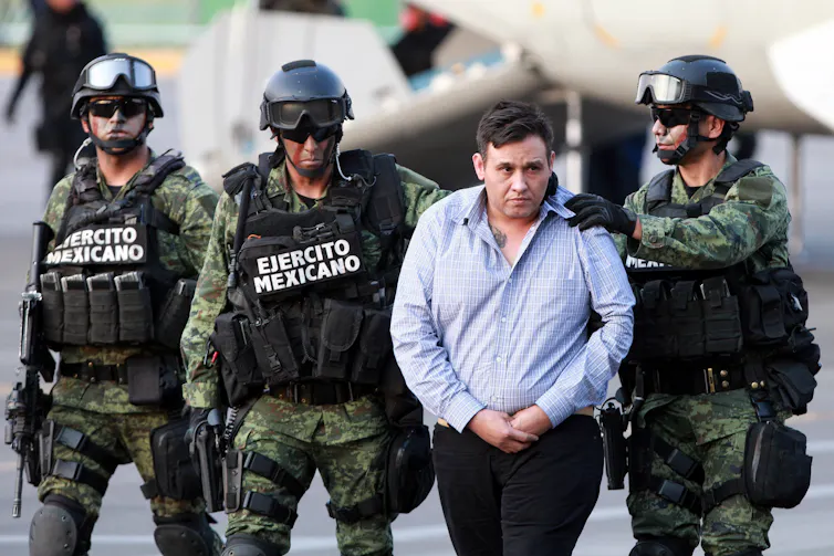 Three armed men escorting a man in a blue shirt and handcuffs.