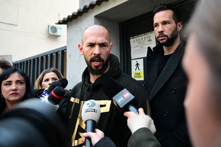 Bald bearded man wearing black sweatshirt surrounded by hands holding microphones.