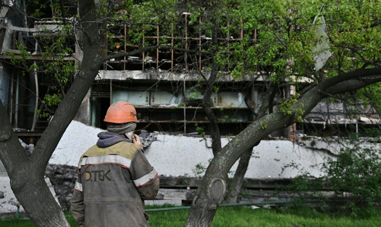 A man wearing an orange hard hat looks at the destroyed shell of a building.