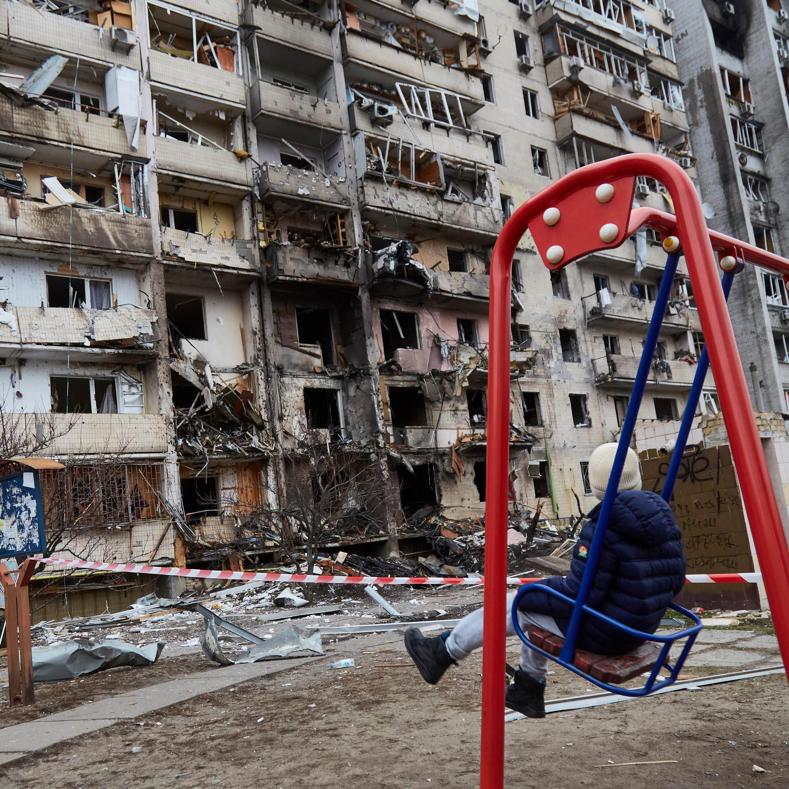 A child on a swing looks at a destroyed building.