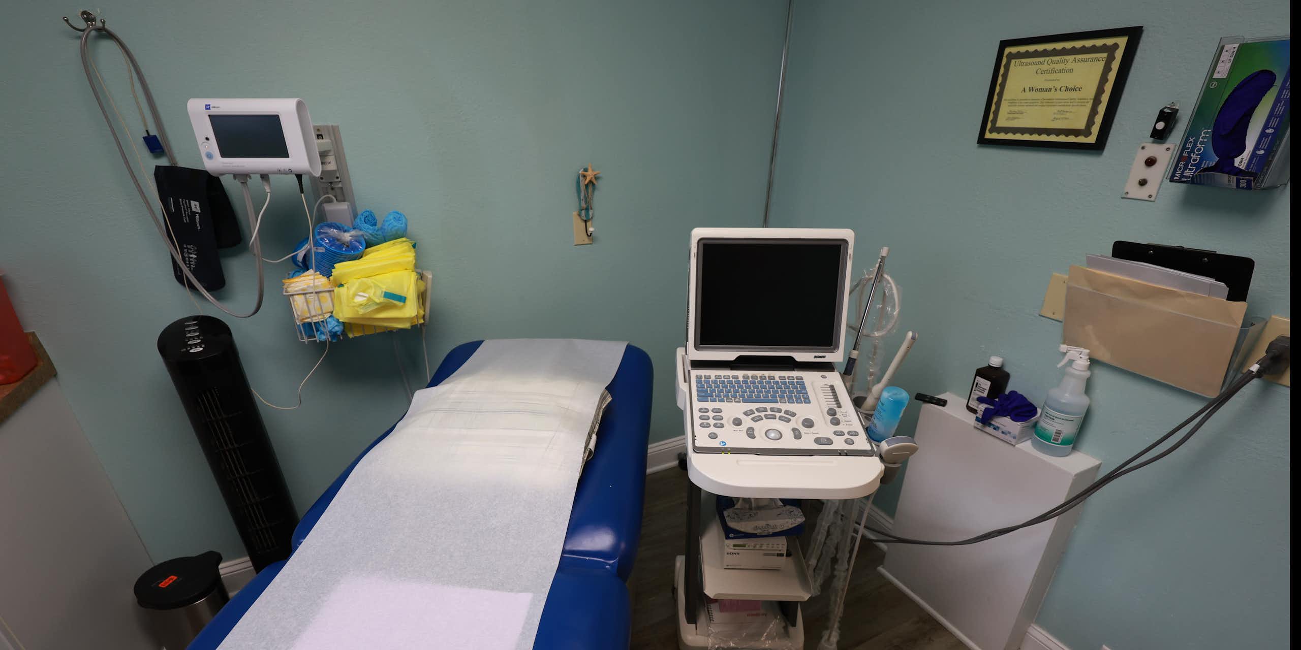 An empty exam room shows a reclining blue doctors chair and medical equipment. 