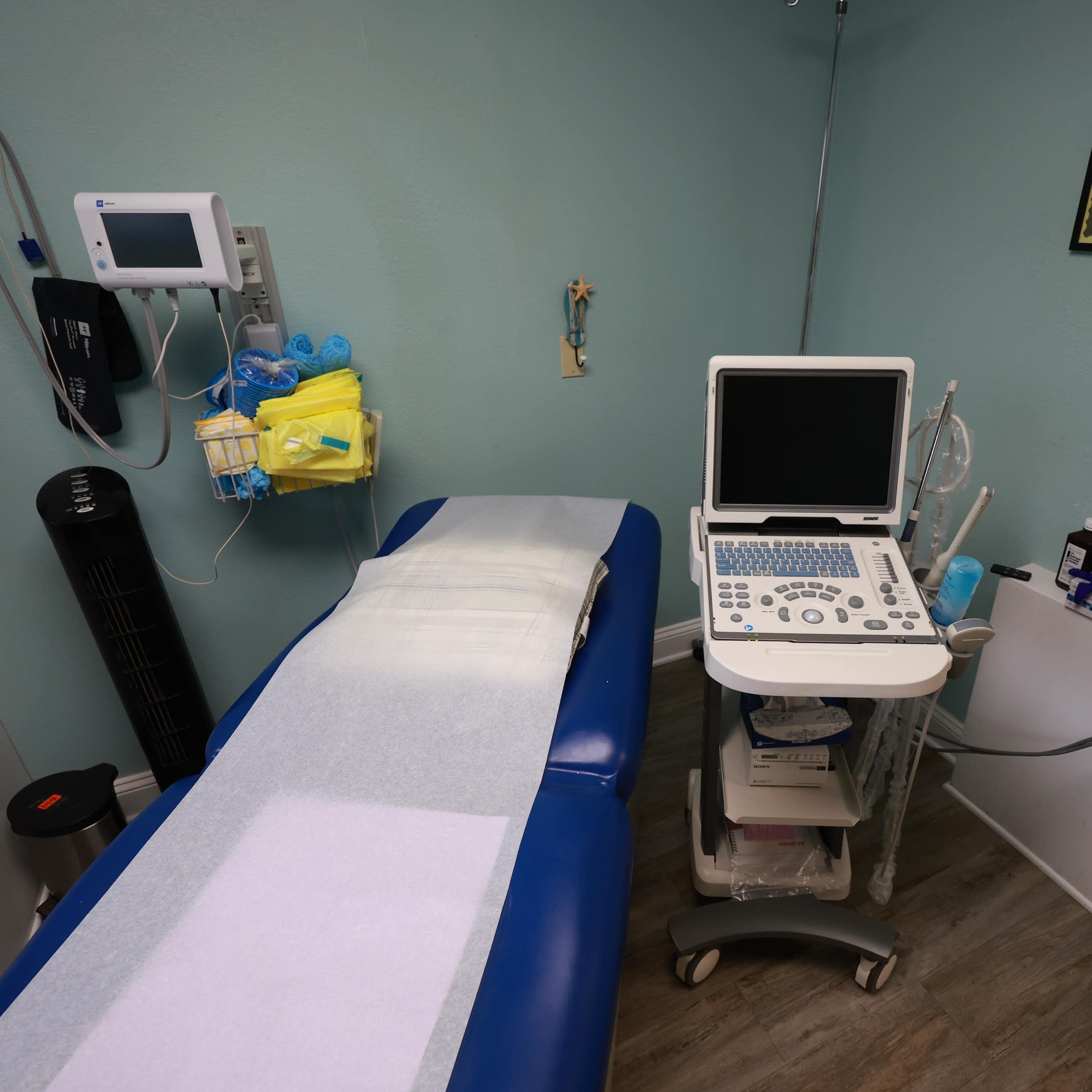 An empty exam room shows a reclining blue doctors chair and medical equipment. 