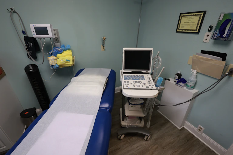 An empty exam room shows a reclining blue doctors chair and medical equipment.