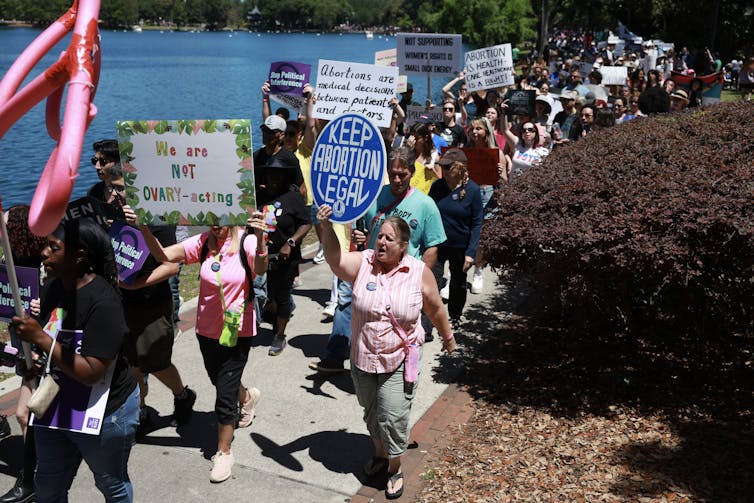 People walk as a group in front of water holding signs that read, “Keep abortion legal.”