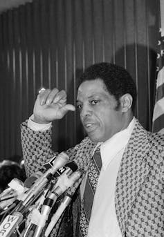 A black-and-white photo of a middle-aged Black man in a suit speaking into a group of microphones