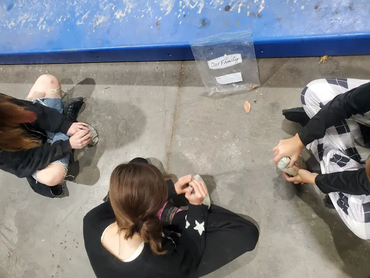Three youths sit crosslegged on the floor, holding rocks and copper nuggets. A copper projectile point rests on the floor between them.