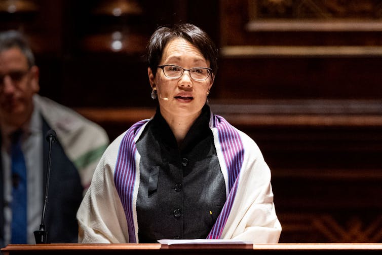 A woman with short black hair and glasses speaks at a podium and wears a white and purple scarf.