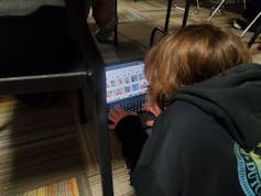 A middle school student lies on a carpeted floor and uses his laptop to search for images. Portraits of Native Americans are visible on the laptop screen.