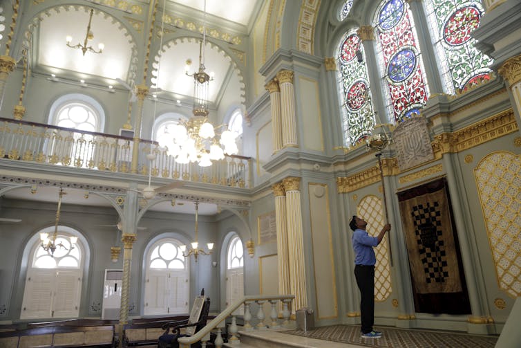 A man in a blue shirt holds up a long tool inside an airy room with stained glass windows.