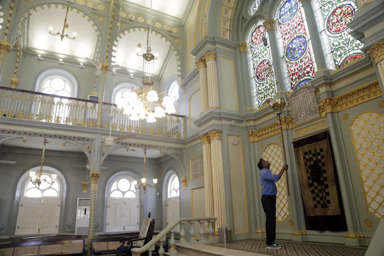 A man in a blue shirt holds up a long tool inside an airy room with stained glass windows.