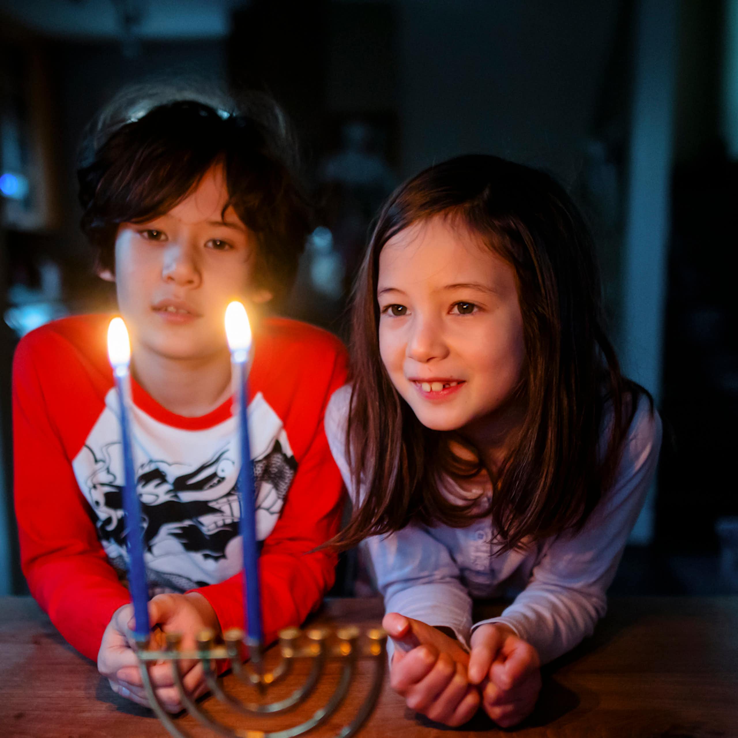 Two children in long-sleeve T-shirts look at two burning candles on a table.