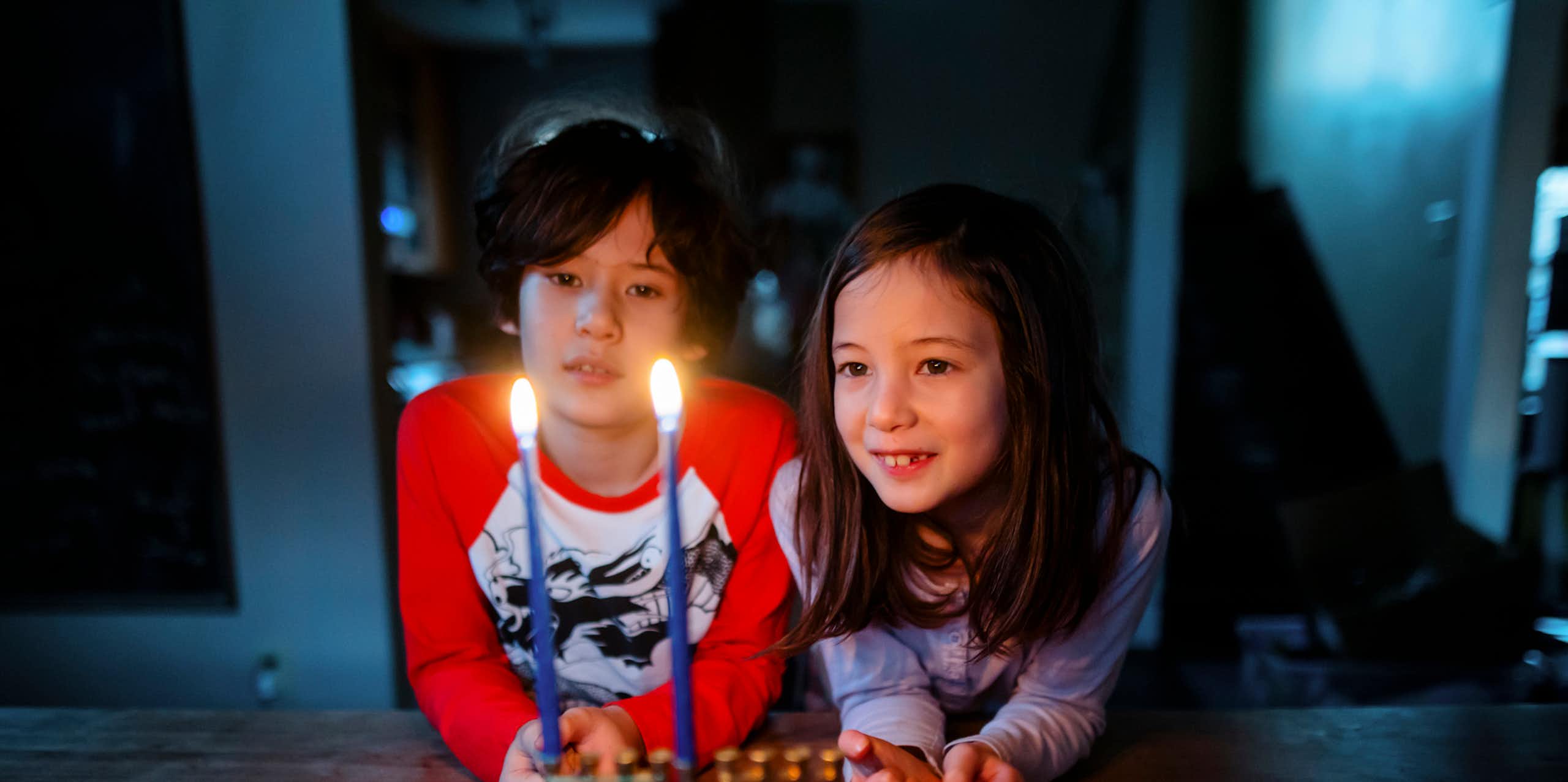 Two children in long-sleeve T-shirts look at two burning candles on a table.