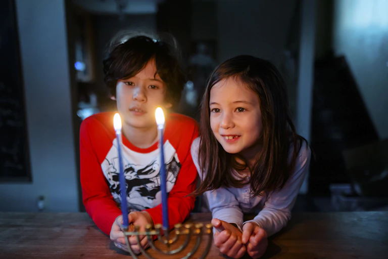 Two children in long-sleeve T-shirts look at two burning candles on a table.