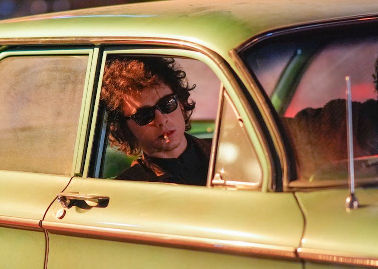 Young man with shaggy curly hair wearing sunglasses and smoking a cigarette, looking out of a car window.