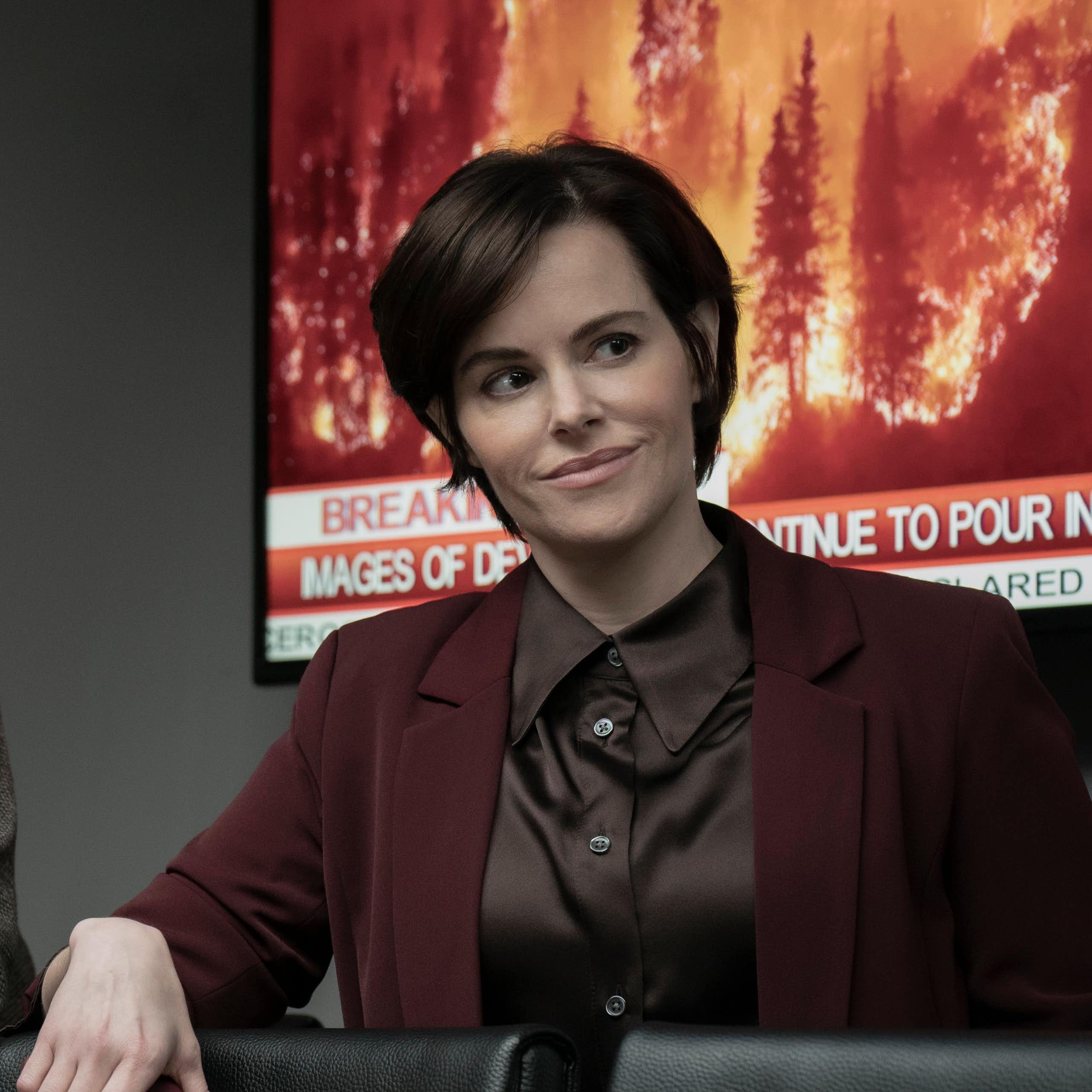 A man and woman in suit jackets with a television image of a forest fire behind them.