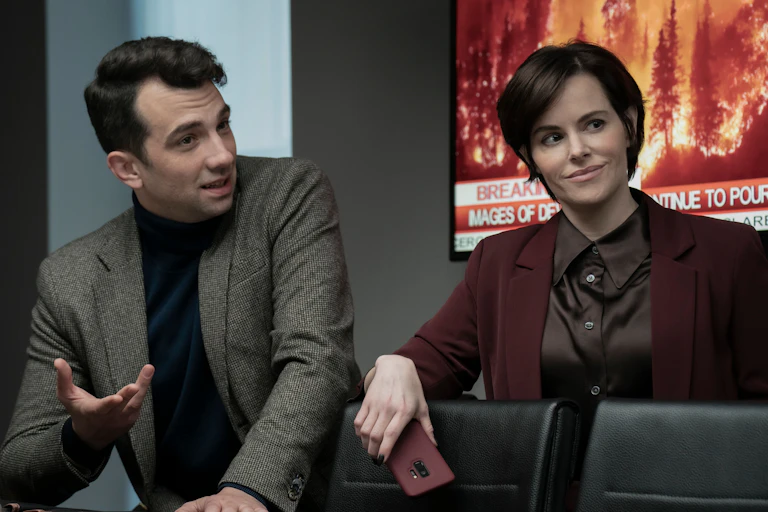 A man and woman in suit jackets with a television image of a forest fire behind them.