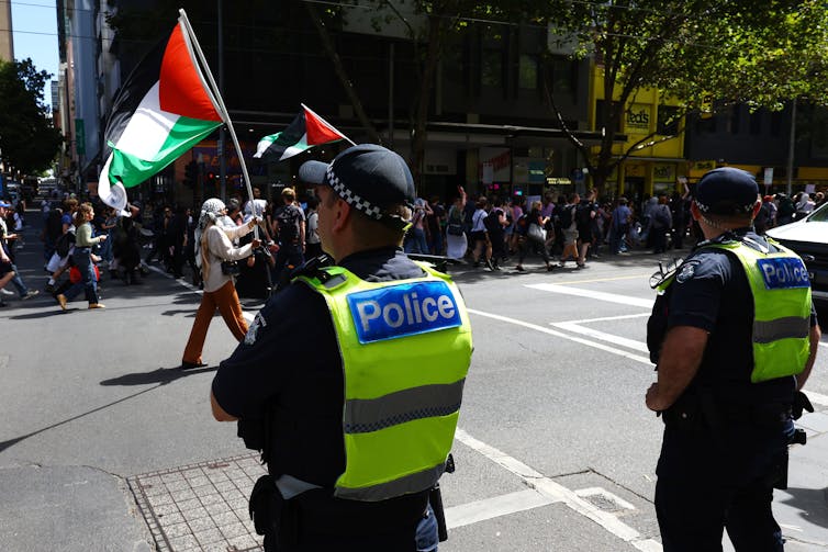 Police officers watch protesters pass them on a street. One protester waves a Palestinian flag.