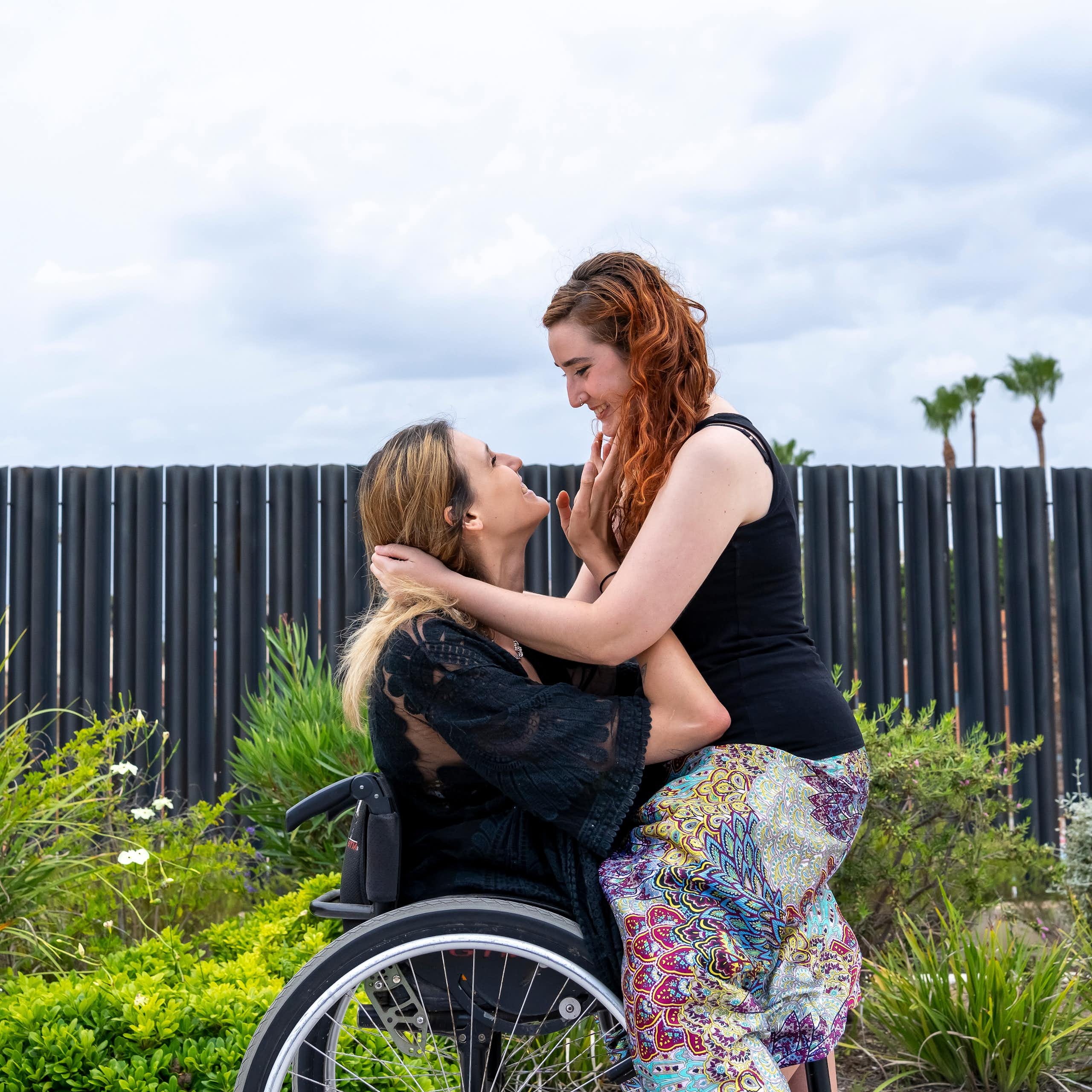 A woman sits on the lap of a woman in a wheelchair. They are smiling and looking lovingly into each other's eyes.