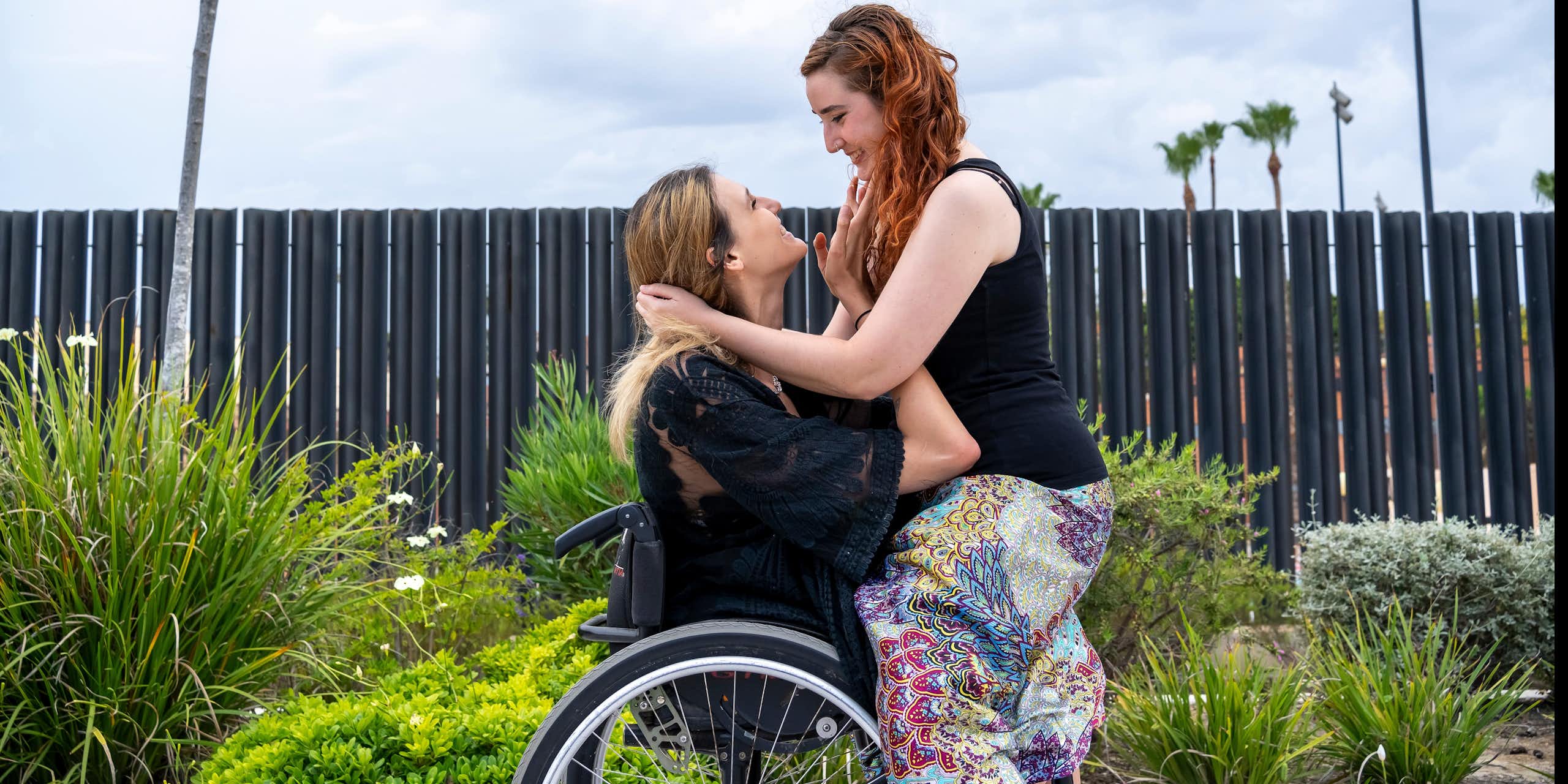 A woman sits on the lap of a woman in a wheelchair. They are smiling and looking lovingly into each other's eyes.