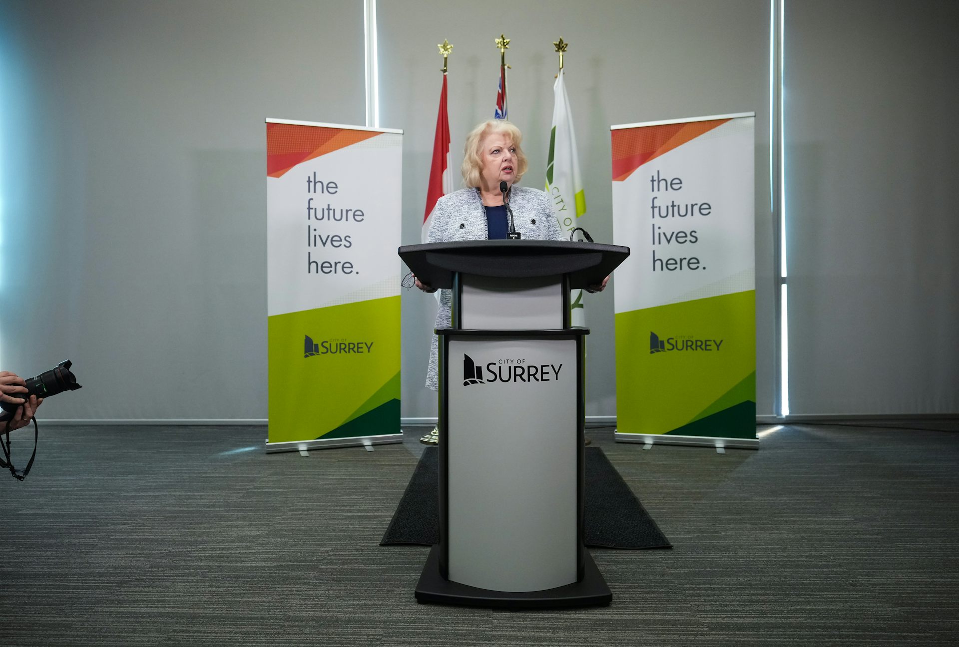 A blonde older woman stands behind a lectern. Signs behind her read The Future Lives Here.