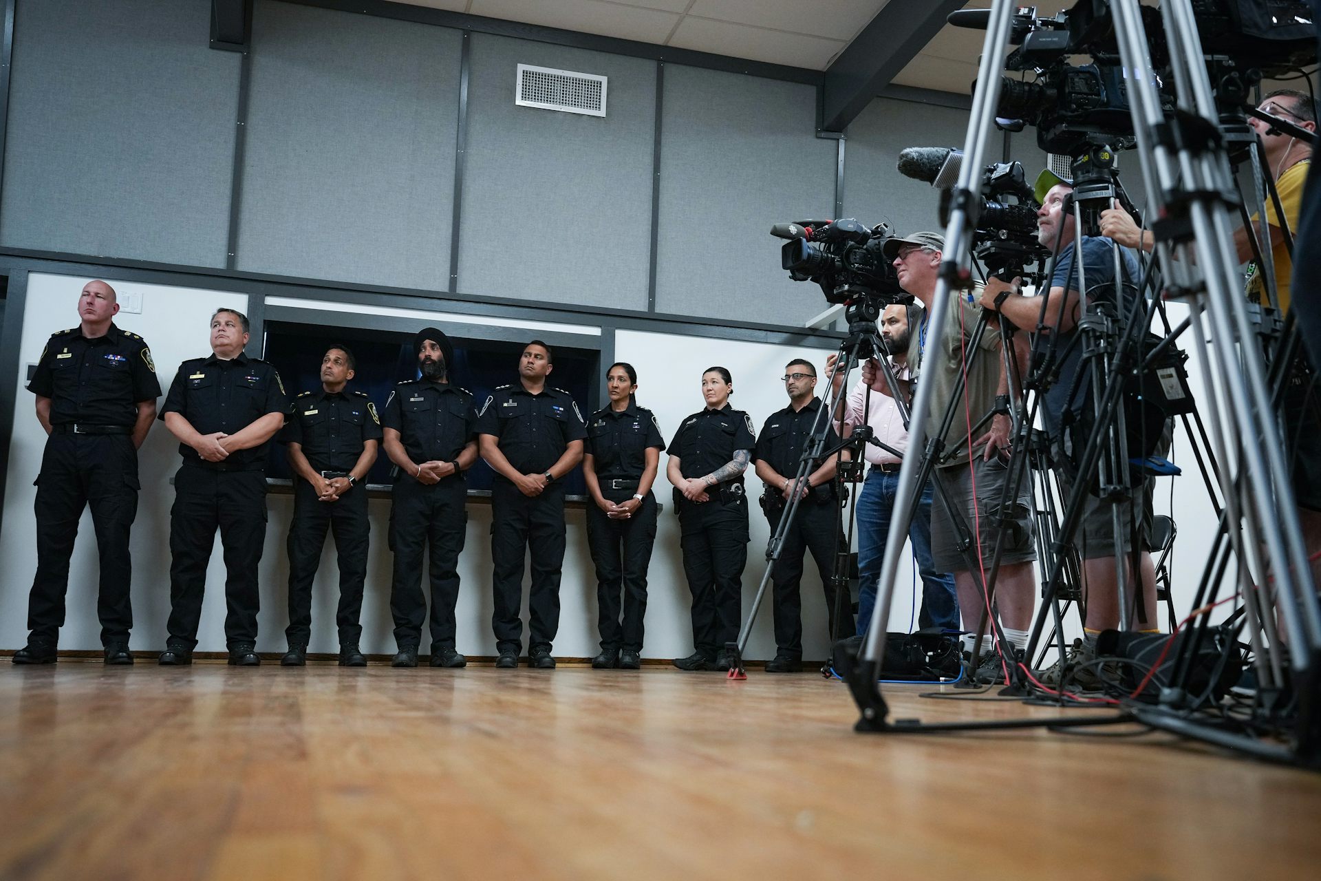 A row of uniformed police officers stand along a walk. A bank of media camera operators are to their left.