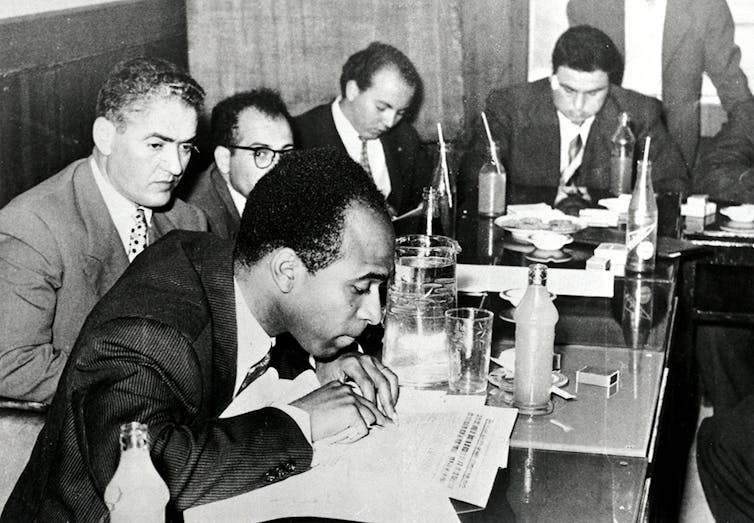 A group of writers, including Fanon, sit around a conference table.