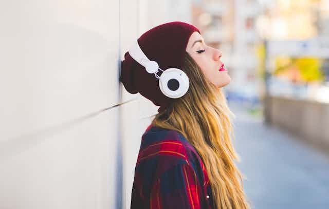 A young woman in a red beanie stands listening to music on headphones.