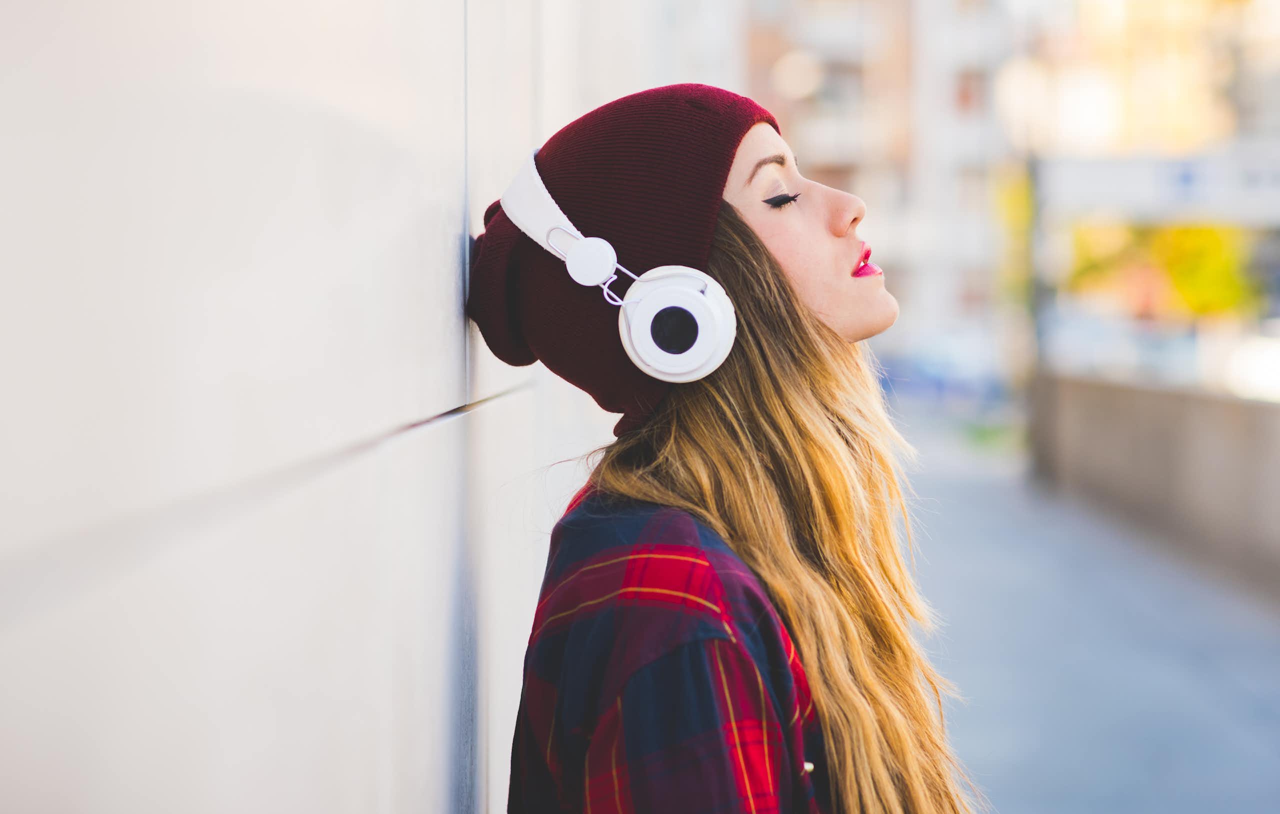 A young woman in a red beanie stands listening to music on headphones.