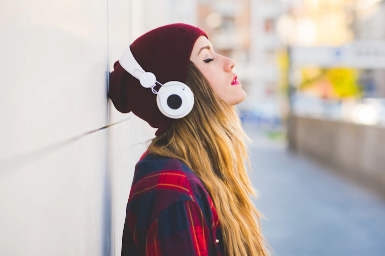 A young woman in a red beanie stands listening to music on headphones.
