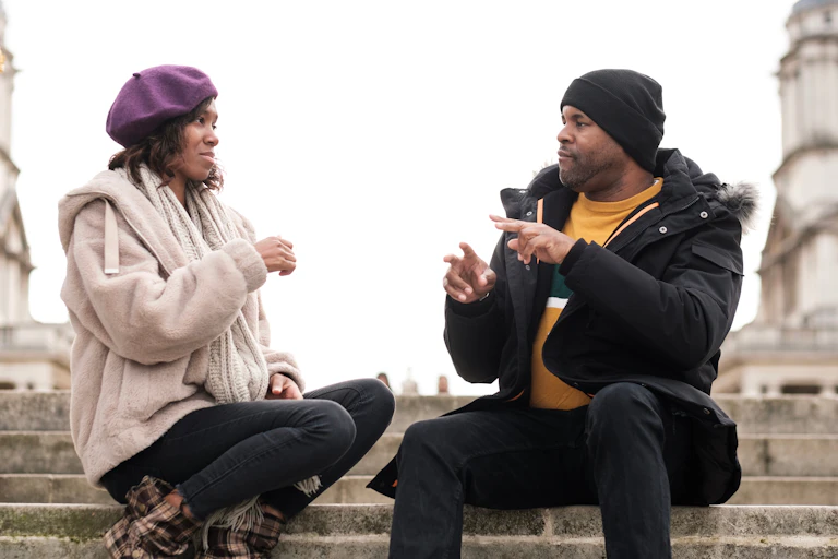 Two friends dressed in winter jackets and hats are sitting on a flight of stairs, talking in sign language