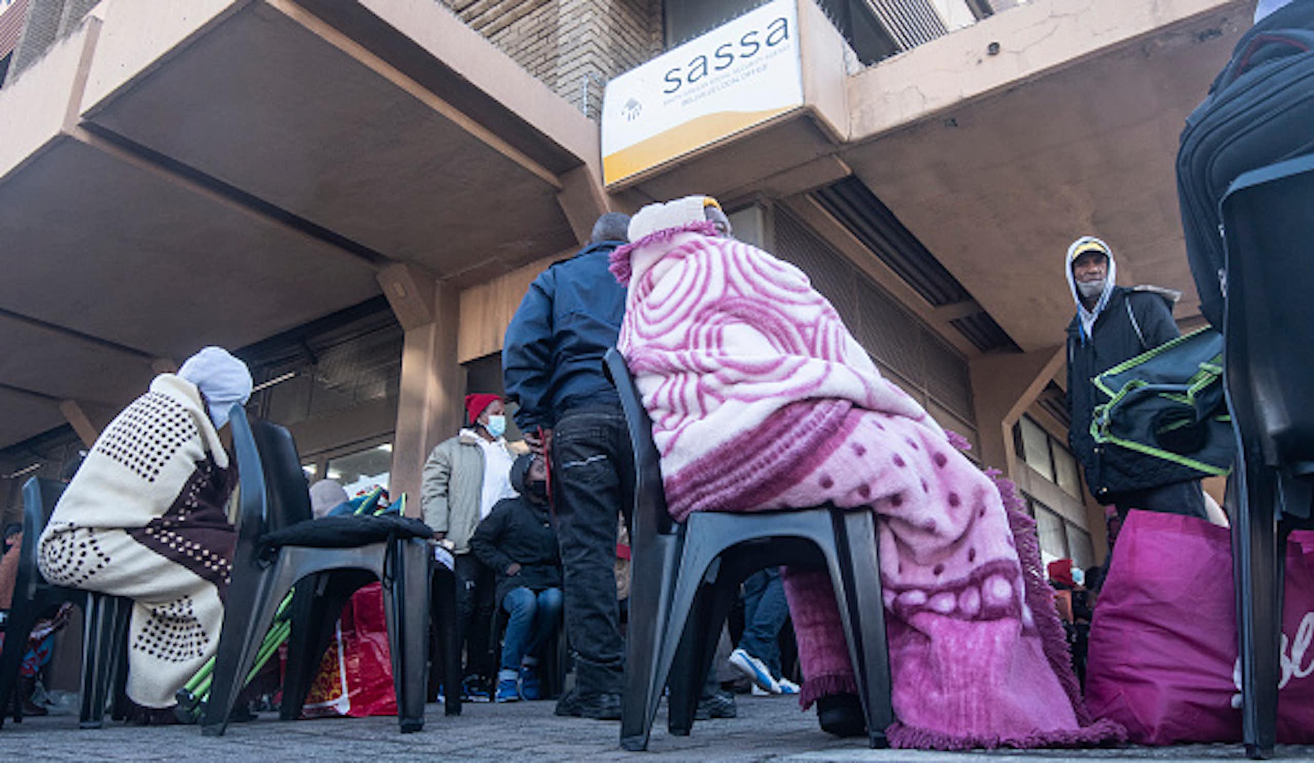 People wearing blankets and covid masks stand or sit in a queue. 