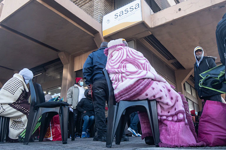People wearing blankets and covid masks stand or sit in a queue.