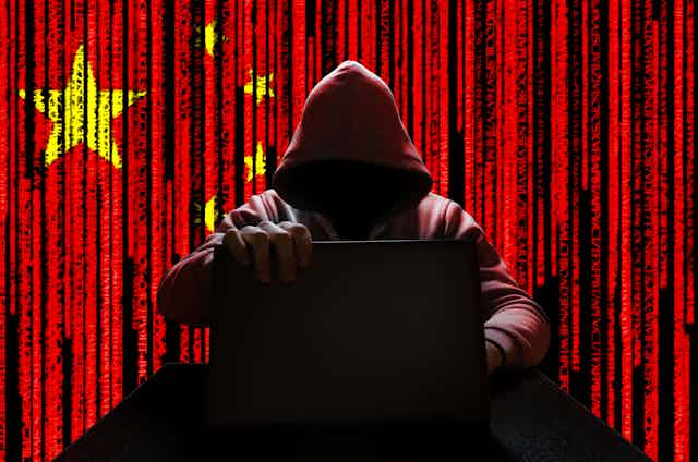 An image of a hooded man holding a laptop against the background of a Chinese flag.