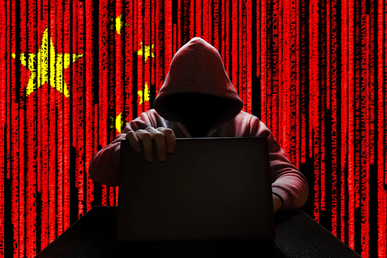 An image of a hooded man holding a laptop against the background of a Chinese flag.