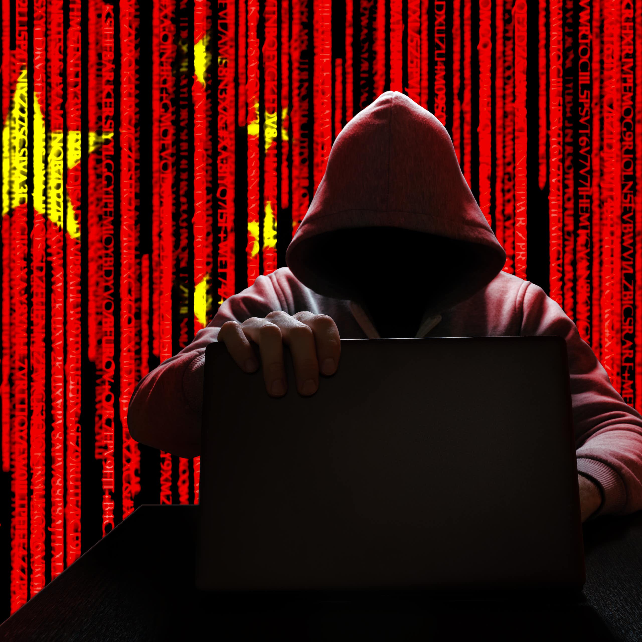An image of a hooded man holding a laptop against the background of a Chinese flag.
