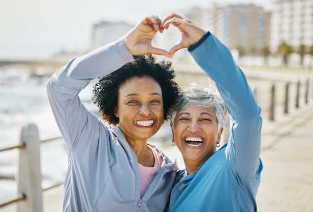 Two women in exercise clothes outdoors, making the heart sign with their hands