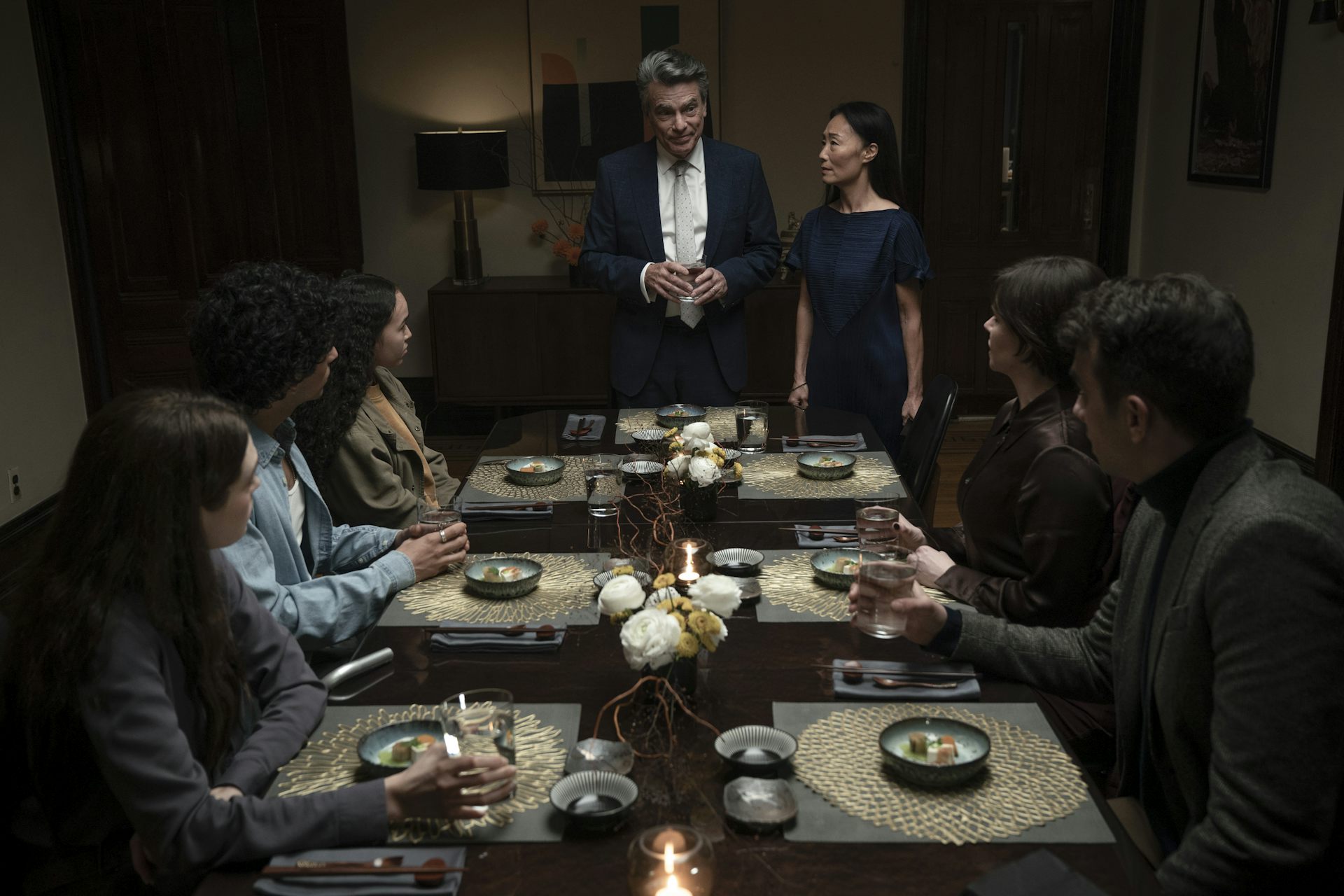 People seen sitting around a dinner table while two people stand at the head of the table.
