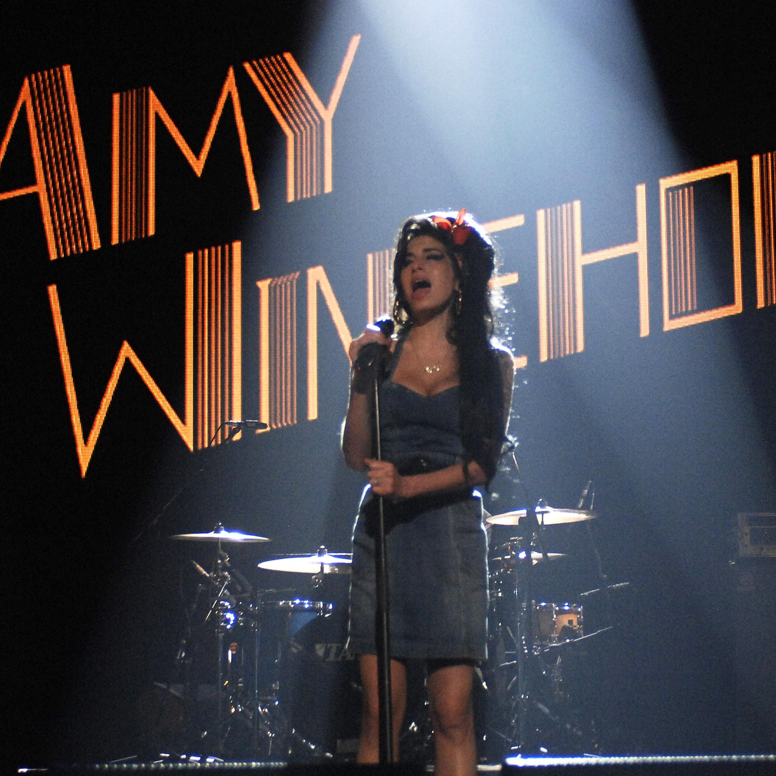 A falecida cantora Amy Winehouse, cujo nome é exibido em luzes, se apresenta em um palco com instrumentos musicais e um guitarrista atrás dela.