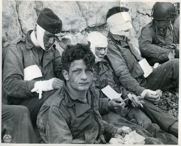 Men in uniforms wearing bandages on their heads, chests and arms sit against a rock wall.