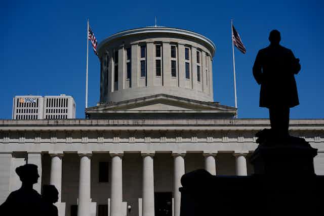 A statue of a man silhouetted against a large gray building with columns and a dome.