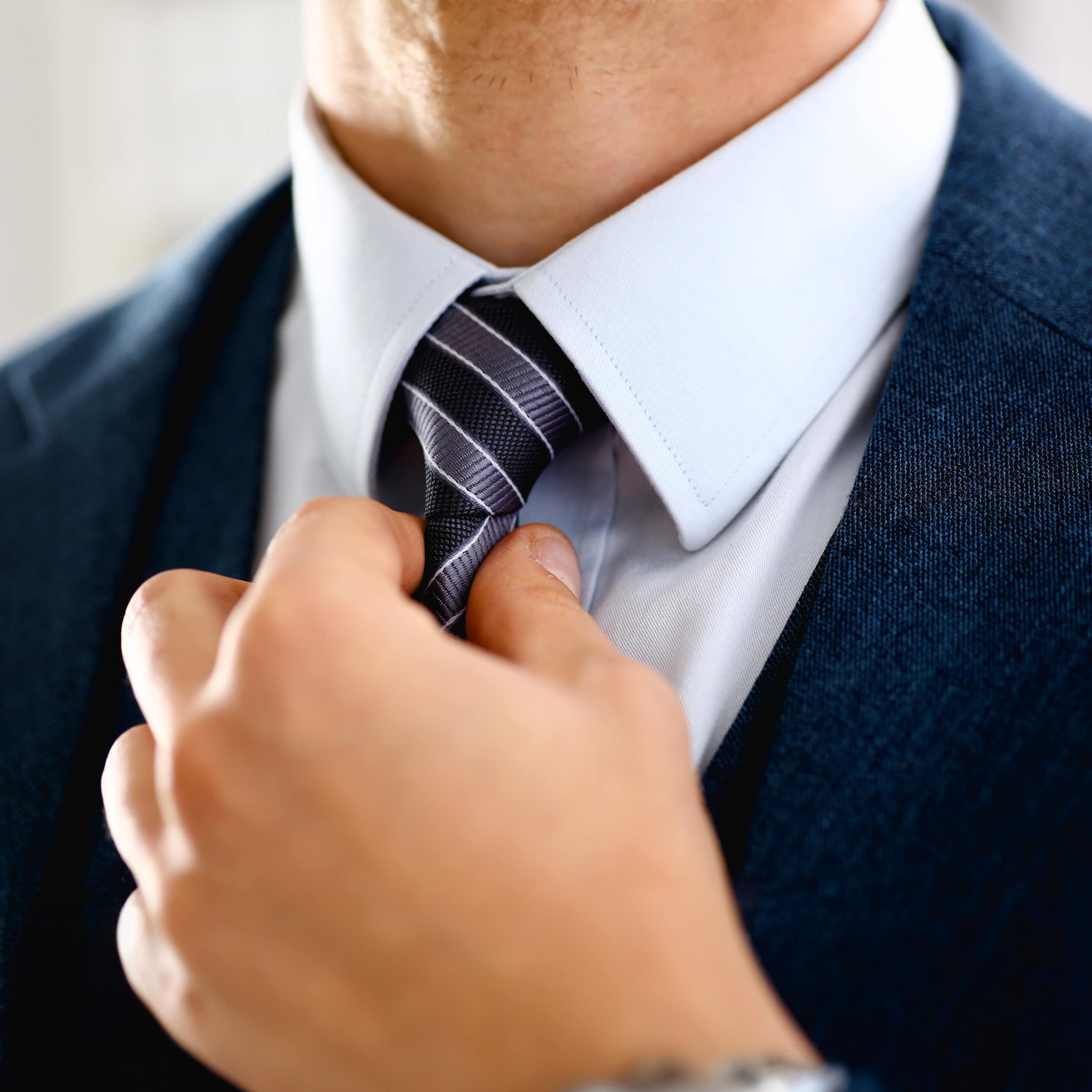 Closeup of a man's hand adjusting a navy blue tie against a light blue collared shirt and navy suit jacket