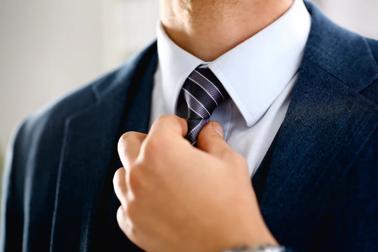 Closeup of a man's hand adjusting a navy blue tie against a light blue collared shirt and navy suit jacket