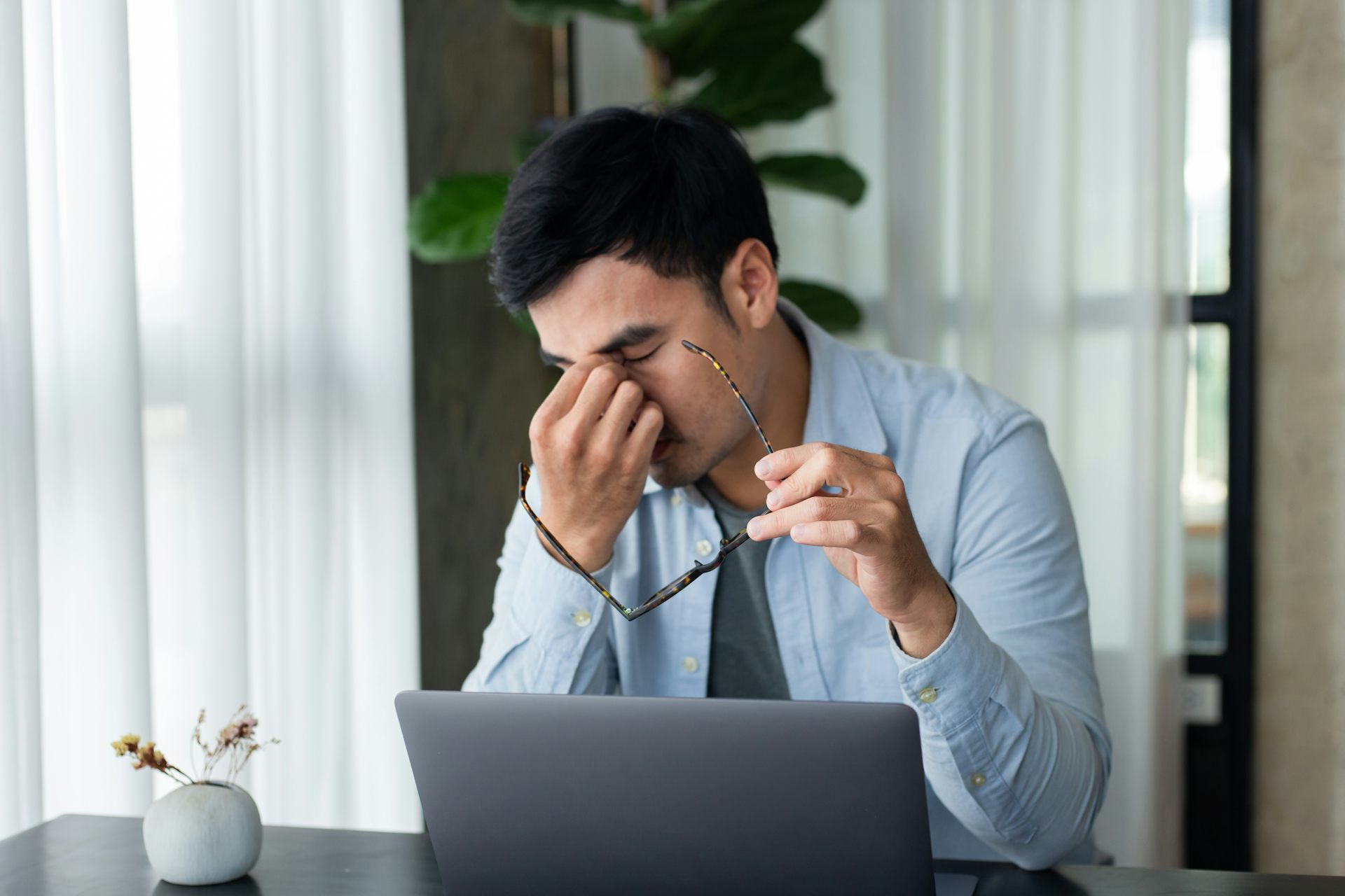 A young, stressed-out-looking man pinches the bridge of his nose with his eyes closed as he sits in front of an open laptop