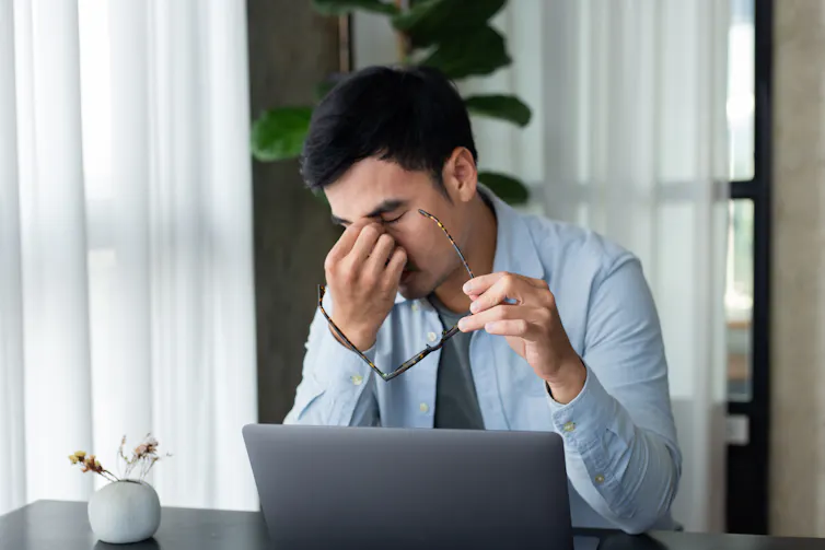 A young, stressed-out-looking man pinches the bridge of his nose with his eyes closed as he sits in front of an open laptop