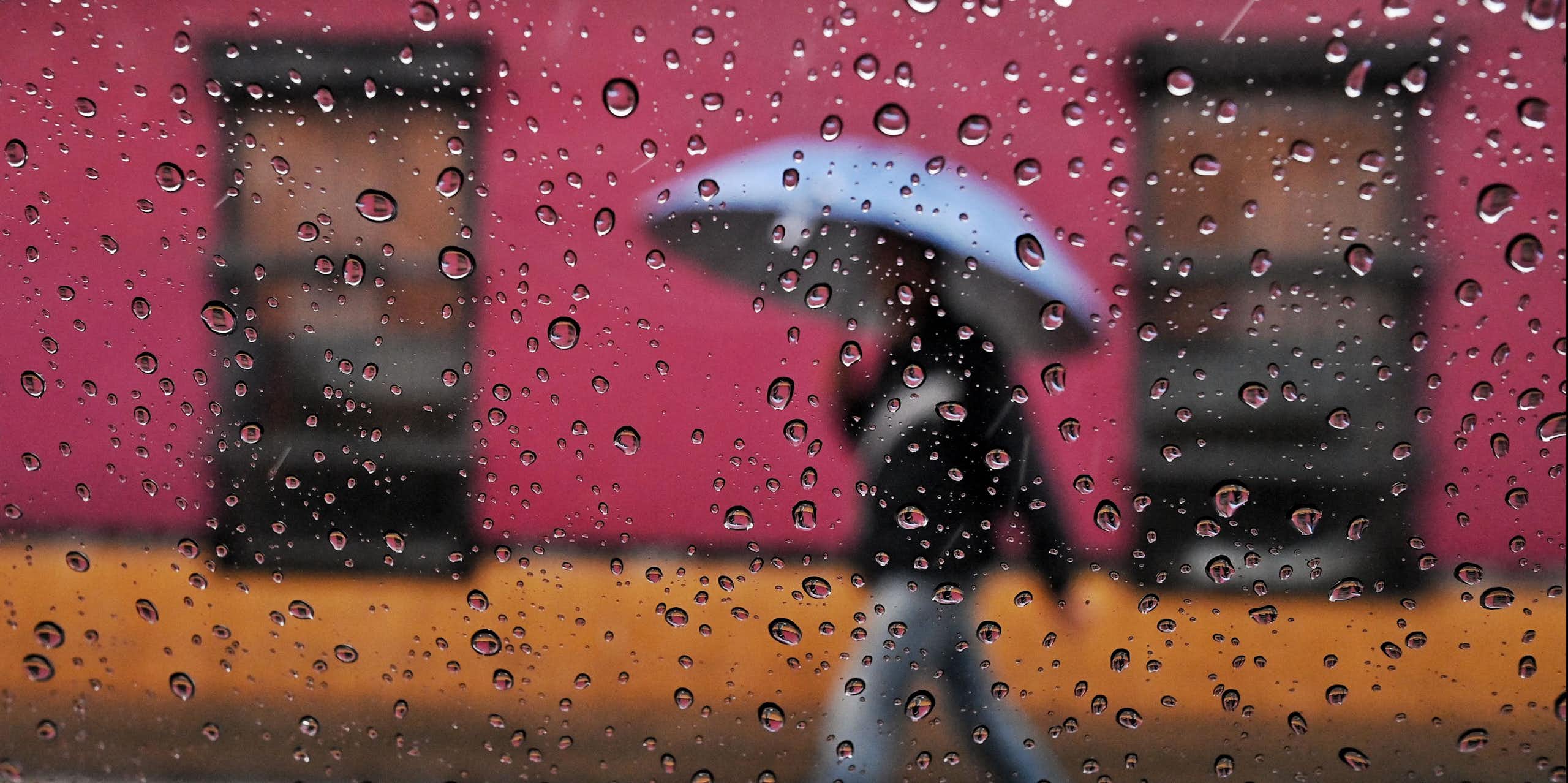 Une personne marche sous la pluie devant des bâtiments aux couleurs vives avec un parapluie. La photo est prise à travers une fenêtre avec des gouttes de pluie sur la vitre.