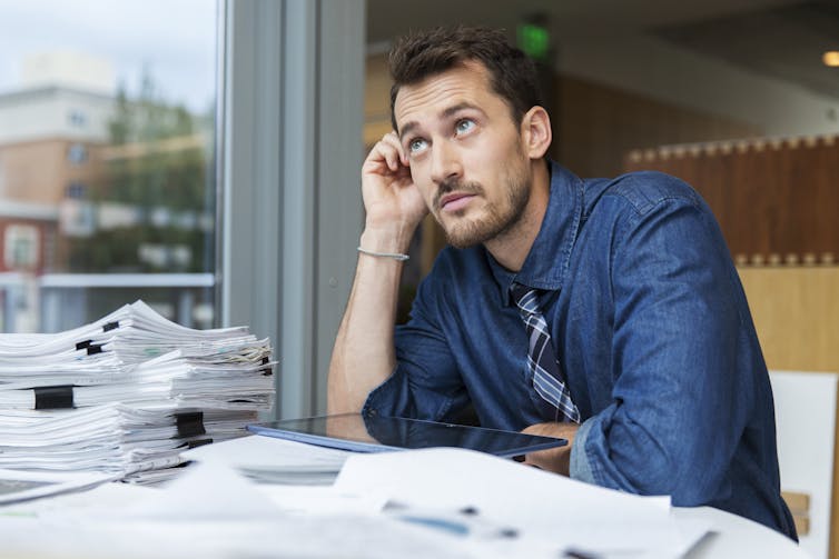 A man appears to daydream while seated at a desk filled with stacks of paper.