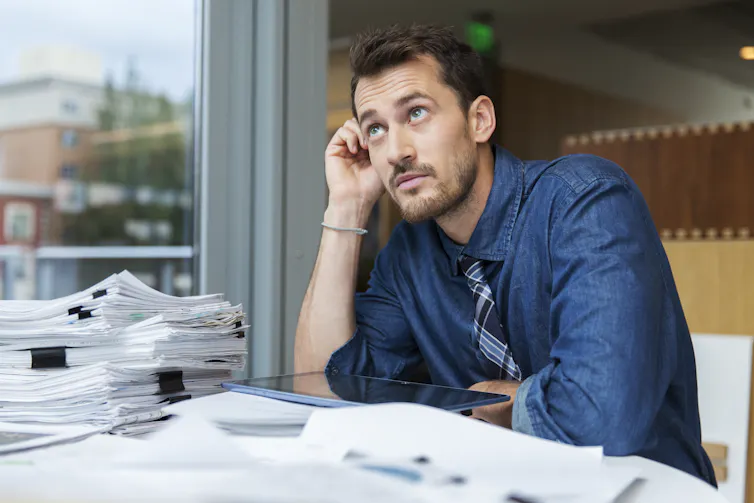 A man appears to daydream while seated at a desk filled with stacks of paper.