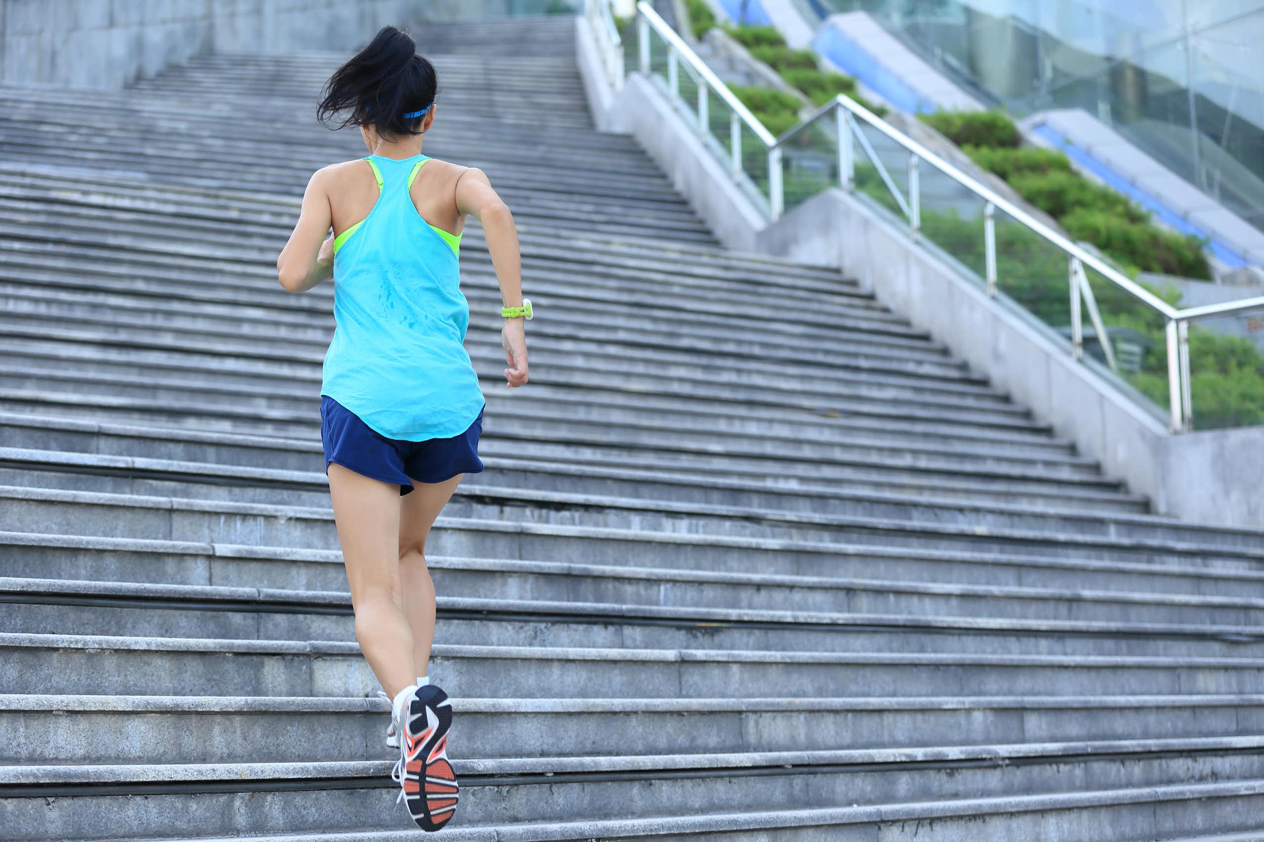 Une jeune femme de dos en tenue de sport monte des escaliers dans un environnement extérieur.